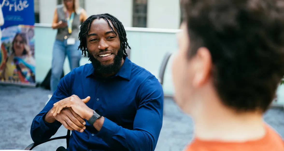 A smartly dressed man in a navy shirt sits comfortably and relaxed, yet engaged in conversation. The setting emphasizes a vibrant, modern environment, reflecting the supportive nature of Aviva’s apprenticeships.
