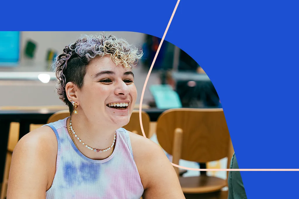 A young woman with short, curly blonde and purple hair, wearing earrings and a necklace, sits in a bright room, smiling. Her joyful demeanour highlights the exciting opportunities of exploring roles through Aviva’s apprenticeships.