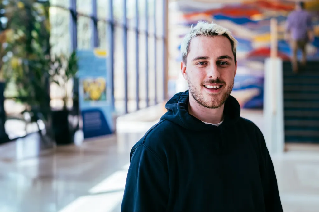 A young man with a beard in a dark hoodie, stands confidently in a modern office foyer with glass walls. The setting reflects the dynamic atmosphere of Aviva’s internships and invites viewers to register now.