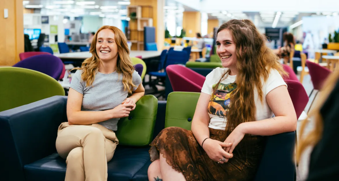 Two confident young women with long hair, dressed in casual clothes, smile brightly while seated on a sofa in a large office space. Their relaxed and positive demeanor reflects the supportive and dynamic environment of Aviva’s graduate programs.