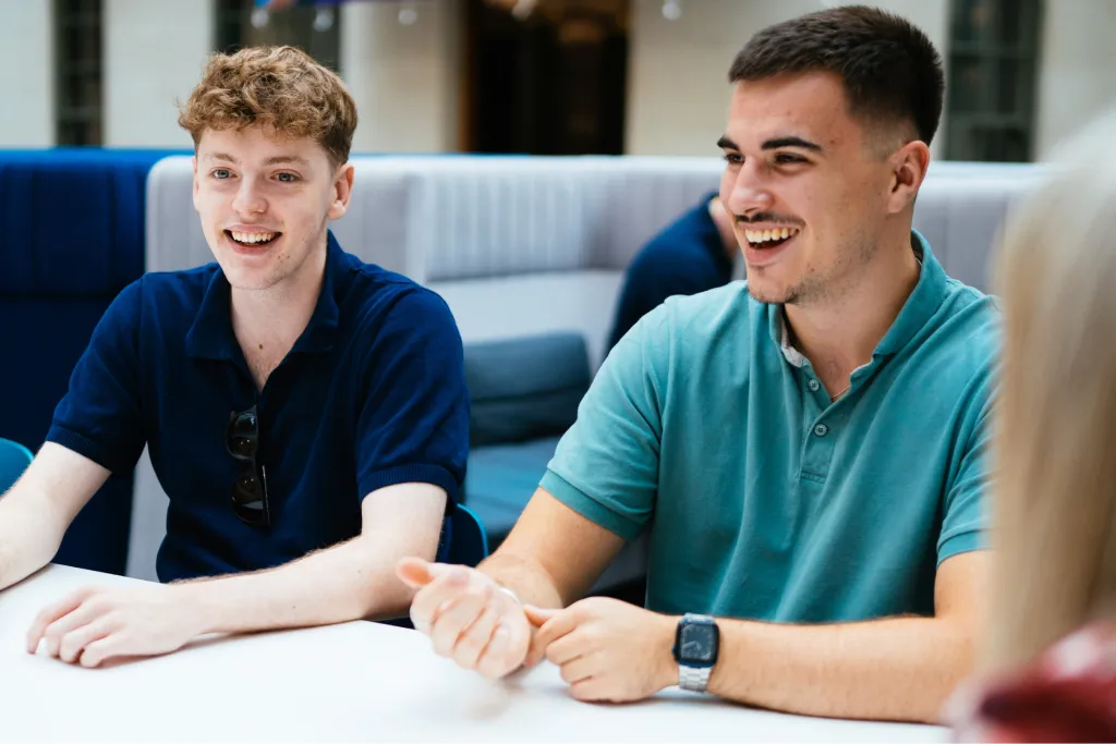 Group of young people happily gathered around a table, engaging and laughing. Two young men in casual shirts are visible, looking joyful. The image reflects the welcoming environment of Aviva's career opportunities for graduates.